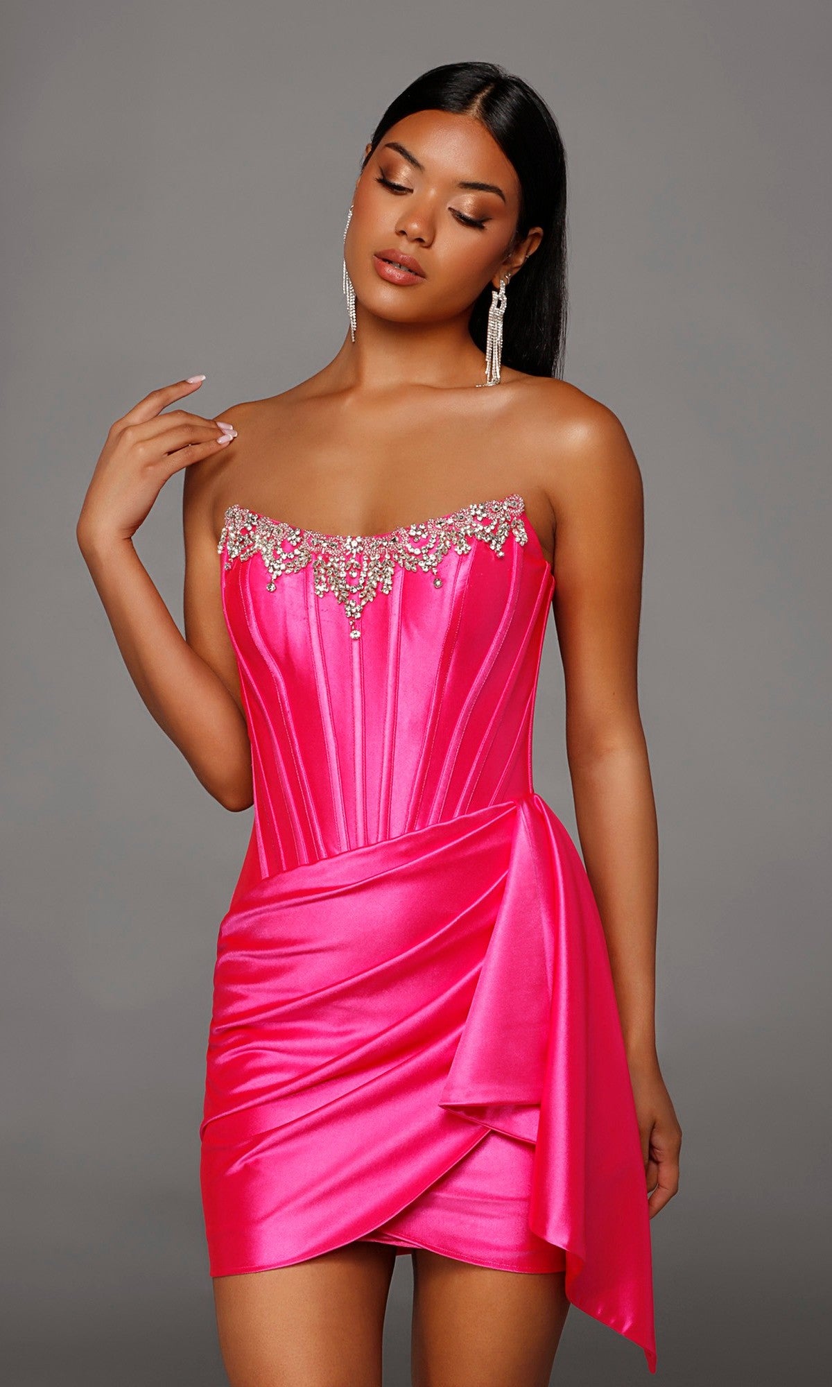 Wearing the Alyce 40207 short homecoming dress in Barbie Pink, a woman stands against a plain gray background. The stretch-satin mini dress features beading along the neckline and a flattering corset bodice.